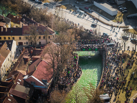 Aerial view of a vibrant green river flowing under a bridge packed with people celebrating St. Patrick's Day, Vilnius, Lithuania.