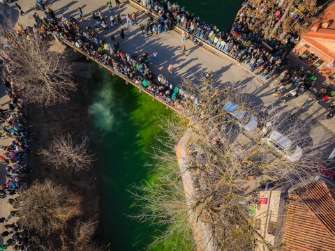 Aerial view of a vibrant green river flowing under a bridge packed with people, casting shadows on a sunny St. Patrick's Day, Vilnius, Lithuania.