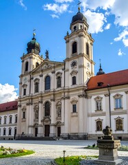Naklejka premium A grand, ornate, beige church with twin towers, red roof, and a blue sky with fluffy clouds. It stands on a cobblestone plaza