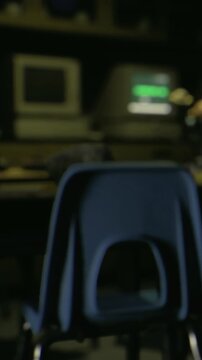 A chair sits in front of a desk with old computers in a classroom. The room has a dark atmosphere with computer screens showing green text. Light comes from the screens.