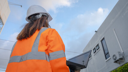 A worker stands outside a building wearing a reflective jacket and helmet. The person looks at a laptop while checking information. It is daytime and the sky is partly cloudy © Happy Photo
