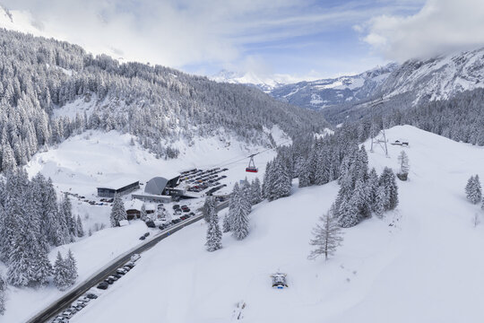 Aerial view of a snowy valley cradling a ski lift station and winding road, surrounded by snow-laden trees, Ormont-Dessus, Vaud, Switzerland.
