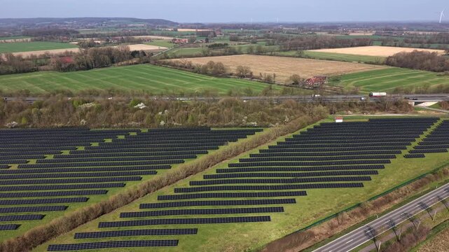 Aerial drone view of large solar farm beside rural highway, with rows of photovoltaic panels across open countryside as traffic passes through peaceful American landscape. Rising wide shot. PA, USA