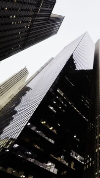 Tall buildings rise high into the sky. The photo shows a contrast between dark and light structures. Clouds fill the sky above the city. People walk below unaware of the view.