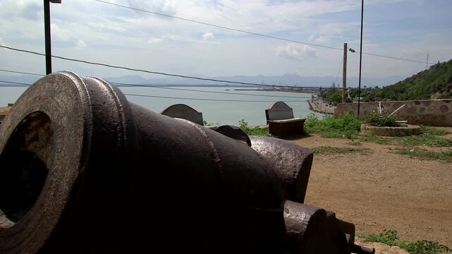 Close-up of rusty colonial cannon at Fort Picolet, Cap-Ha&iuml;tien, Haiti: Barrel aimed at bay with stone benches and walls, distant mountains under blue sky with wires.