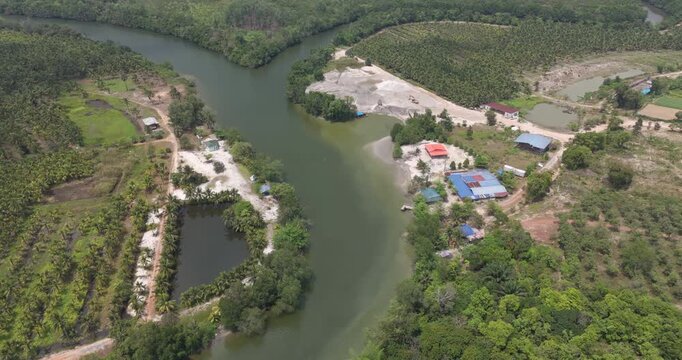 Forward aerial view of a winding river revealing a sand dredging site, palm plantations, and colorful buildings beside an aquaculture pond.