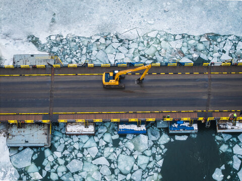 Aerial view of a yellow excavator amidst the fractured ice floes surrounding a bridge, a stark contrast against the dark waters, Gdansk, Poland.