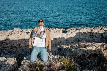 Fototapeta premium Stylish man in casual outfit standing on rocky coast, enjoying sunlight. Modern lifestyle, confidence, travel mood and outdoor freedom in scenic seaside environment.