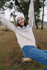 Fototapeta premium Young woman on a swing outdoors, wearing a cozy sweater and beanie, smiling joyfully in a park with trees, grass and soft daylight today