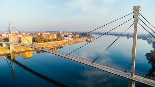 Aerial view of the pedestrian bridge gracefully stretches over the still, reflective Sava River, connecting the city under a crisp, clear sky, Sremska Mitrovica, Vojvodina, Serbia.