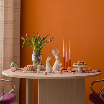 Minimalist Easter brunch setup featuring a modern wooden table, vibrant tulips, and soft pastel decorations against a bold orange wall.