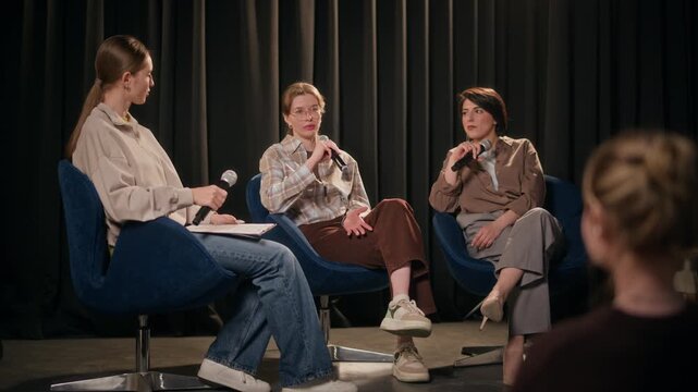Full length shot of three young adult female comedians performing in conversational panel style format on stage sharing stories during live show at comedy venue