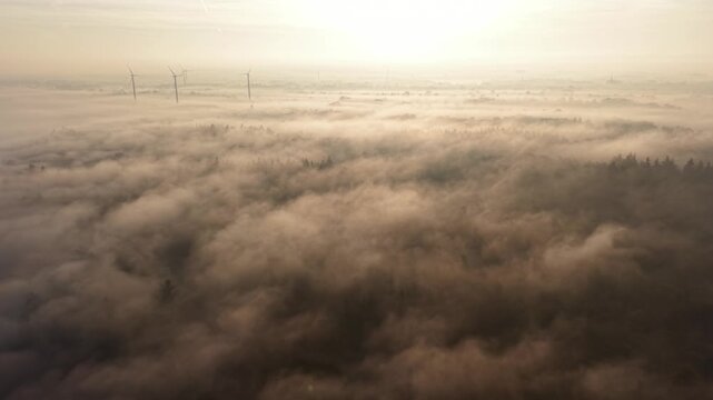 Aerial view of a misty river at golden sunrise. Thick pink fog over rural fields with wind turbines on the horizon. Cinematic morning landscape, peaceful nature and clean energy concept.