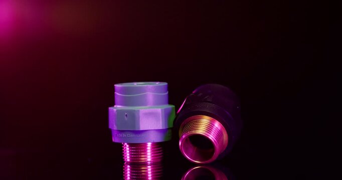 Two industrial plastic pipe fittings with metal threads standing on a dark surface with red lighting