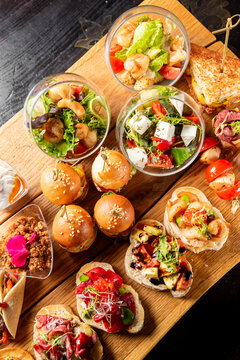 Top view of a diverse catering spread on a wooden board featuring mini burgers, bruschettas with various toppings, fresh salads in glasses, and gourmet appetizers for a party or event.