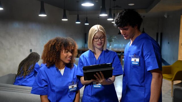 Group of multiethnic medical students wearing blue scrubs collaborating in a hospital hallway, analyzing a patient's medical records and discussing treatment options on a digital tablet