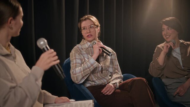 Rack focus shot of three young adult female comedians sharing funny stories performing in comedy panel show on stage of stand up comedy club