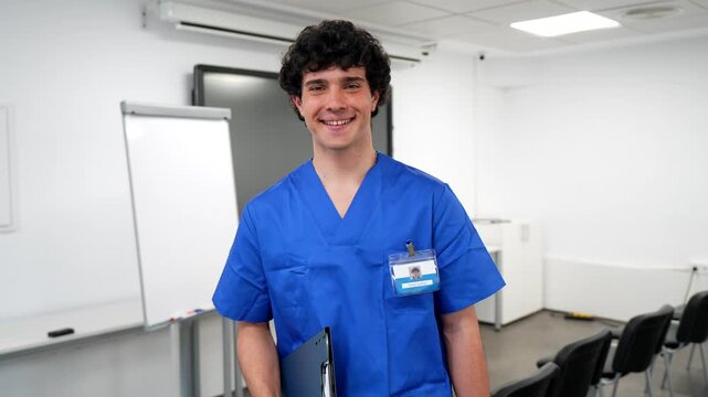 Portrait of a young male nurse or medical student wearing blue scrubs and holding a clipboard, smiling confidently at the camera inside a modern hospital classroom or conference room