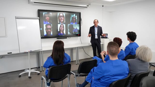 Group of medical students in blue scrubs attending a hybrid conference with a professor presenting in a modern classroom, connecting with remote colleagues via a large interactive digital screen