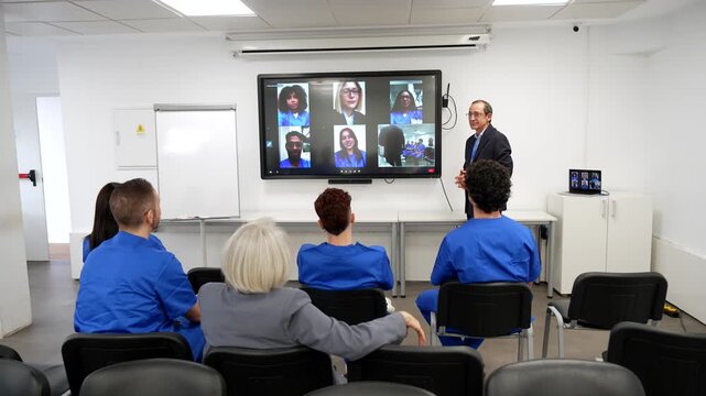 Medical staff in blue scrubs attending a hybrid seminar in a modern hospital classroom, with a speaker presenting a video conference call on a large interactive whiteboard to remote participants
