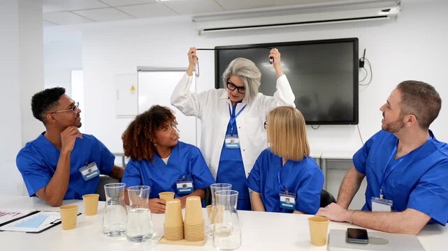 Mature female professor in a white coat holding a stethoscope and lecturing a multicultural group of young medical interns wearing blue scrubs during a training seminar in a modern hospital room