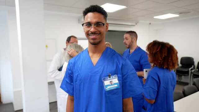 Portrait of a young african american male nurse smiling at the camera. Wearing blue scrubs and an id badge. With his multicultural medical team discussing in the background of a hospital meeting room