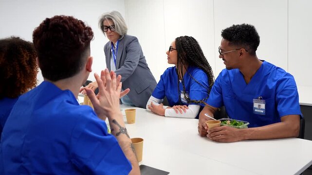 Multiracial group of young nurses and medical interns in blue scrubs listening to a mature female doctor during a meeting in the hospital canteen, having a healthy lunch and discussing work