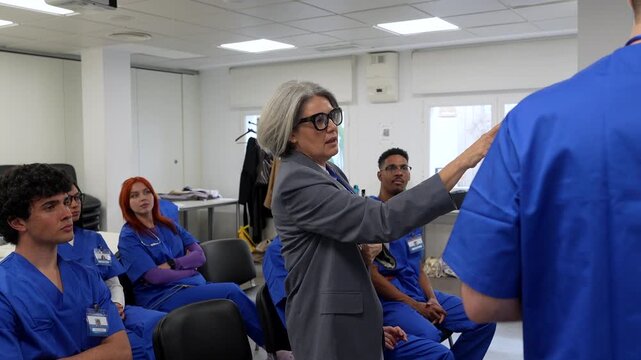 Mature female professor lecturing a group of young, diverse medical students in blue scrubs during a university lesson, explaining complex healthcare topics in a hospital training room