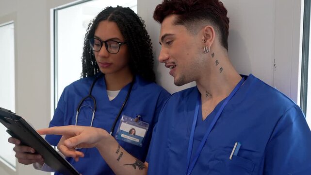 Two young multicultural medical professionals wearing blue scrubs discussing a patient's medical records on a clipboard in a bright hospital corridor, demonstrating teamwork and collaboration