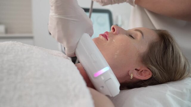 A beautician in gloves performs a non-invasive radiofrequency lifting procedure on a woman's face and neck in a beauty clinic. The client is relaxed during the skin tightening treatment.