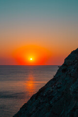 Obraz premium A bright orange sunset over the sea with a dark cliff silhouette in the background. Sunset sky, ocean horizon, coastal nature.