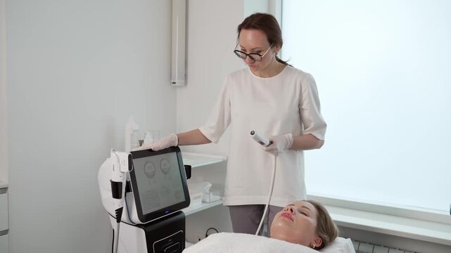 A professional cosmetologist in a modern clinic performs a non surgical facelift procedure on a female client. The beautician uses advanced hardware cosmetology for skin rejuvenation and tightening.