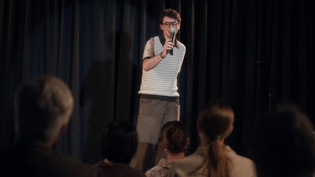 Crowd level view of cheerful young adult male comedian interacting with audience giving answer based improvisation during standup comedy show on stage against black curtains