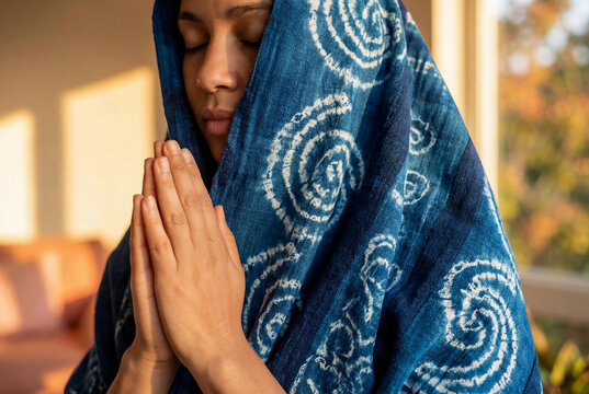 Peaceful young woman praying with closed eyes wearing blue indigo Adire fabric head wrap in warm sunset light for mindfulness and spirituality concept