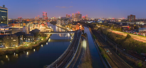 Obraz premium Aerial panorama at night showing the River Irwell, Bridgewater Canal, and Metrolink tracks leading to the Manchester skyscraper skyline, UK.