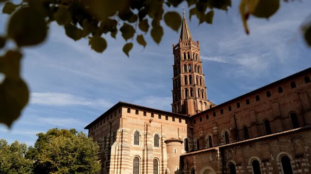 Low angle view of the iconic Basilica of Saint-Sernin in Toulouse showing its tall octagonal bell tower and warm pink brick Romanesque facade glowing in sunlight against a blue wispy sky