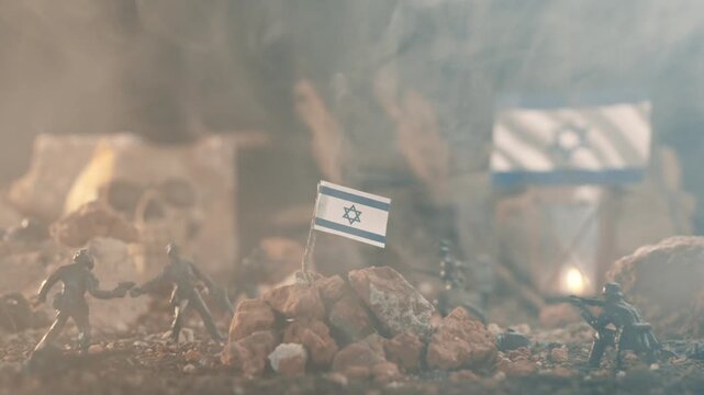 Diorama of a warzone with toy soldiers. An israeli flag stands on a pile of rocks amidst smoke, fire, and a human skull representing loss