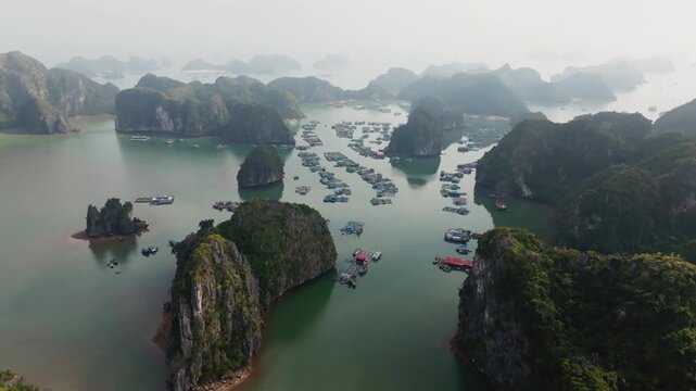 Cat Ba Vietnam drone forward flight over dense floating village and fish cages between karst islets, misty distance adding depth.