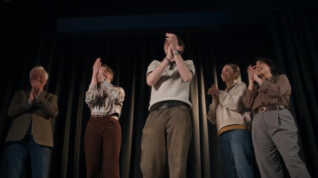 Low angle shot of multigenerational crew of stand up comedians throwing up script sheets while giving final bow on stage of comedy venue