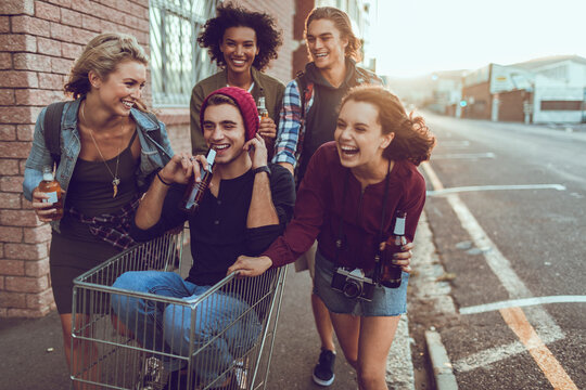 Group of young adults laughing with drinks, pushing a shopping cart on an urban street at sunset