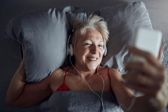 Smiling senior woman using smartphone with headphones in bed at home