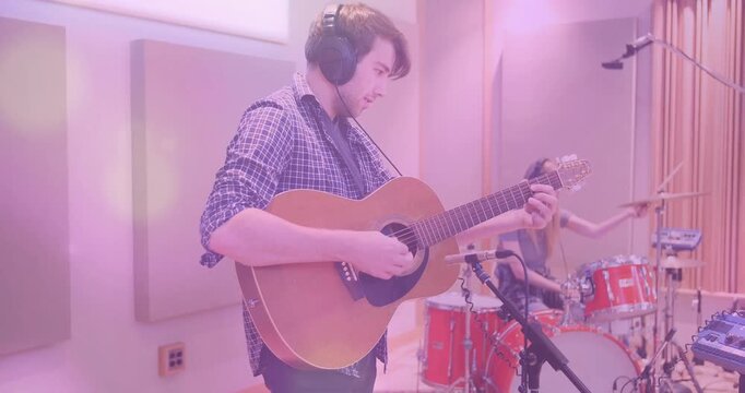 Male guitarist pressing frets and strumming chords in studio while drummer hitting drums for rhythm