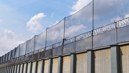 Obraz premium High-security prison concrete wall fence with netting and barbed wire under bright cloudy blue sky (1)