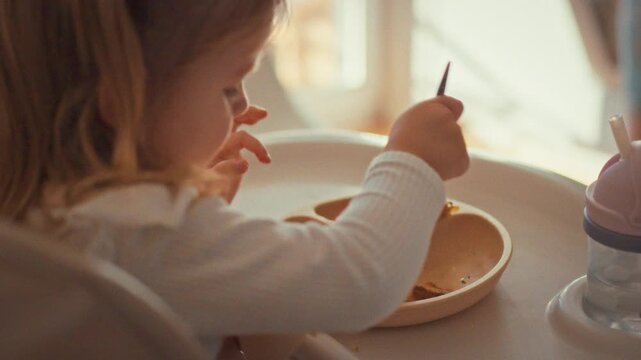 Sitting securely baby chair, little girl wears protective apron as she enjoys meal. With fork in hand, she practices independent eating, mastering daily skill self-feeding amidst messy-friendly setup