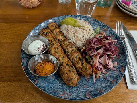 Chicken kofta kebabs served with rice, yogurt sauce, harissa, pickled vegetables, and pickles on a blue patterned plate near the Skylon in Greater London, United Kingdom.