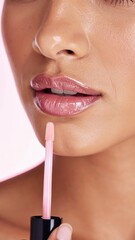 Naklejka premium A detailed close-up image of a woman applying glossy pink lipstick, highlighting smooth skin and lip texture against a soft pink backdrop.