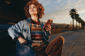 Fototapeta premium A woman in a rainbow sweater and denim jacket smiles while holding a smartphone leaning against a car at sunset on a palm tree lined road. Lifestyle and technology concept.