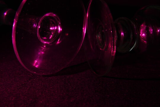 Copas de vidrio para tragos,  transparentes con vibrantes reflejos de luz rosa sobre una superficie oscura abstracta, con  elegantes curvas  iluminadas por una llamativa luz magenta