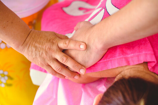 A horizontal top-down close-up shot of a therapist's hands performing a manual massage or osteopathic manipulation on a patient's shoulder. The patient is covered with a bright pink towel.