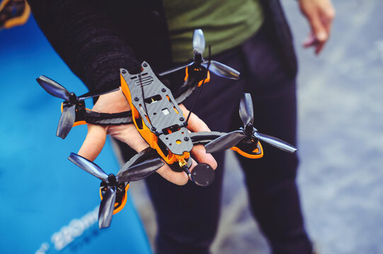 A horizontal shot of a fully assembled FPV racing drone with propellers, held by a person. The drone has a black carbon fiber body and bright orange accents.